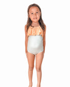 Ein kleines Kind namens Ava steht barfuß und lächelt in die Kamera. Sie trägt einen ärmellosen, hellen Ava Mädchen Badeanzug von Main Design mit verstellbaren Trägern und einem Rüschenoberteil in einem Pfirsichton. Der Hintergrund ist schlicht weiß.