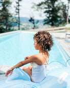 Ein kleines Kind mit lockigem Haar sitzt auf einer aufblasbaren Schwimmhilfe am Rand eines klaren, blauen Schwimmbeckens. Das Kind trägt einen Ava Mädchen Badeanzug von Main Design, komplett mit verstellbarem Träger, und blickt in die Ferne. Im Hintergrund sind Bäume und eine malerische Aussicht auf sanfte Hügel und Berge zu sehen.