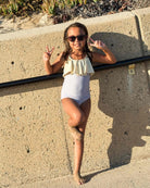 Ein Mädchen trägt den Ava Mädchen Badeanzug von Main Design mit verstellbaren Trägern und steht barfuß an einer sonnenbeschienenen Steinmauer. Sie lächelt, zeigt das Peace-Zeichen und genießt die entspannte Strandkulisse.