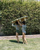 Zwei Personen in passenden grünen Kaia Mädchen Badeanzügen von Main Design tragen ein Surfbrett über ihren Köpfen, während sie an einem sonnigen Tag auf Gras in Richtung einer Hecke laufen.