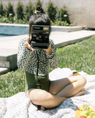 Der Kaia Mädchen Badeanzug von Main Design mit UPF 50+: Ein Mädchen sitzt im schnell trocknenden Anzug am Pool auf einer Decke, hält eine Polaroid-Kamera, neben ihrem Obst, im Hintergrund sind Pool und Grünpflanzen.