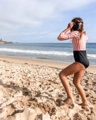 Ein Kind in einem gestreiften und schwarzen Laila Mädchen-Badeanzug von Main Design aus recycelten Materialien steht an einem Sandstrand in der Nähe des Ozeans und blickt an einem teilweise bewölkten Tag auf das Wasser.