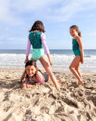 Drei lächelnde Mädchen genießen einen Sandstrand im Laila Mädchen Badeanzug von Main Design aus recycelten Materialien. Zwei stehen, während eines im Sand liegt, während sich unter einem wolkenverhangenen Himmel die Wellen nähern.