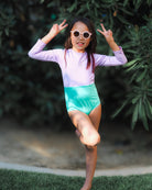 Ein Mädchen trägt den Laila Mädchen Badeanzug von Main Design mit UPF 50+ und runden Sonnenbrillen, steht auf einem Bein, zeigt das Peace-Zeichen mit beiden Händen – im Hintergrund grünes Laub.