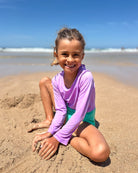 Ein lächelndes Kind, das den Main Design Laila Mädchen Badeanzug und die blaugrünen Shorts trägt, genießt den Strand, das Meer und den blauen Himmel - und bleibt dabei mit UPF 50+ geschützt.