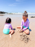 Zwei kleine Kinder im passenden Laila Mädchen Badeanzug von Main Design mit UPF 50+ spielen im Sand an einem sonnigen Strand, mit Wellen und Felsen im Hintergrund.