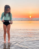 Ein junges Mädchen im Laila Mädchen Badeanzug von Main Design steht bei Sonnenuntergang barfuß im seichten Strandwasser und hält sanft einen Fisch. Der UPF 50+ Schutz und das nachhaltige Design zeigen ihre Sorge um die Natur und den Komfort.