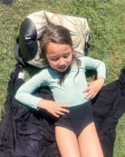 Ein junges Mädchen in einem Laila Mädchen Badeanzug von Main Design liegt auf einem schwarzen Handtuch im Gras, stützt den Kopf auf eine gemusterte Tasche und genießt die Sonne im Freien.