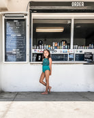 Ein junges Mädchen im blaugrünen Laila Mädchen-Badeanzug von Main Design, hergestellt aus recycelten Materialien mit Sonnenschutzfaktor 50+, steht barfuß vor einer Snackbar mit einer schwarz-weißen Speisekarte und Getränkedosen im Fenster.