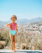 Ein kleines Kind trägt den Lena Mädchen Badeanzug von Main Design, einen roten und hellblauen UPF 50+-Badeanzug, und posiert im Freien auf einem Stein mit einer verschwommenen Stadtlandschaft und Bergen unter einem klaren blauen Himmel im Hintergrund.