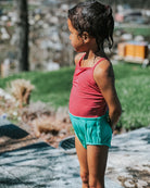 Ein kleines Kind im Main Design Lena Mädchen Badeanzug - mit rotem UPF 50+ Oberteil und blaugrünen Shorts - steht mit nassen Haaren auf einem sonnenbeschienenen, grasbewachsenen Bürgersteig und genießt den Tag in diesem nachhaltigen Badeanzug.