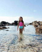 Ein junges Mädchen steht im klaren, flachen Wasser zwischen Felsen am Strand und trägt den Lena Mädchen Badeanzug von Main Design aus recycelten Materialien in Pink und Blau. Ein heller Himmel und eine ferne Küstenlinie vervollständigen die Szene.