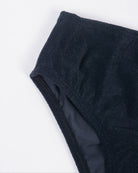 Nahaufnahme des Mia Mädchen Bikiniunterteils von Main Design aus strukturiertem schwarzem Stoff. Das Bild konzentriert sich auf die Ecke und hebt das Material und die Nahtdetails aus recycelten Materialien vor einem schlichten hellen Hintergrund hervor.