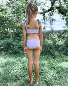 Ein junges Mädchen mit Pferdeschwanz steht barfuß im Gras und trägt den Mia Mädchen Bikini reversibel von Main Design in hellem Lila. Sie befindet sich im Freien zwischen Bäumen und Grünpflanzen und schaut von der Kamera weg.