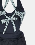 Nahaufnahme des Milly Mädchen Badeanzugs von Main Design in Schwarz mit abstrakt gepunkteten weißen und schwarzen Trägern, die im Rücken zu einer Schleife gebunden werden, vor einem schlichten weißen Hintergrund.