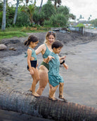 Eine Frau und zwei kleine Kinder in Main Design Milly Mädchen Badeanzug, alle mit sandigen Beinen, balancieren auf einem Treibholzstamm an einem tropischen Strand mit Palmen und üppigem Grün im Hintergrund.