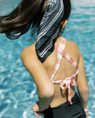 Ein Mädchen mit langen braunen Haaren in einem Pferdeschwanz und einem schwarz-weißen Halstuch steht an einem Pool und trägt einen rosa-weißen Milly Mädchen Badeanzug von Main Design mit gekreuztem Rücken und Schleife. Sie schaut von der Kamera weg.