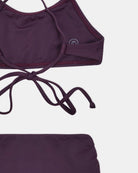 Eine Nahaufnahme des Nalu Mädchen Bikini von Main Design in dunklem Lila, hergestellt aus recycelten Materialien, auf weißem Hintergrund. Das Oberteil hat dünne Träger, die in der Mitte gebunden werden, hier mit einem Teil des passenden Unterteils.