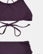 Eine Nahaufnahme des Nalu Mädchen Bikini von Main Design in dunklem Lila, hergestellt aus recycelten Materialien, mit einem Oberteil im Neckholder-Stil mit Bindeband im Rücken, einem passenden gerüschten Unterteil und UPF 50+ Sonnenschutz auf weißem Hintergrund.