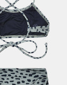 Eine Nahaufnahme des Nalu Mädchen Bikini von Main Design, hergestellt aus recycelten Materialien, mit einem dunklen Oberteil mit dünnen gekreuzten Trägern und Bindeband, einem hellen Unterteil und einem unregelmäßigen schwarzen Punktmuster.