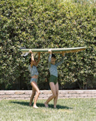 Zwei Mädchen tragen barfuß das Surfbrett über das Gras, bekleidet mit passenden Nalu Mädchen Bikinis von Main Design; im Hintergrund sind eine grüne Hecke und eine Steinumrandung zu sehen.