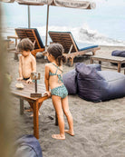 Zwei kleine Kinder in dem Nalu Mädchen Bikini von Main Design stehen an einem Tisch mit einem 74er Schild an einem Sandstrand, umgeben von Sitzsäcken und Sonnenschirmen - ideal für Kinderspaß mit UPF 50+ Sonnenschutz.