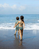 Zwei kleine Kinder, eines in einem Main Design Nalu Mädchen Bikini und das andere in Shorts und Hemd, laufen barfuß über den Sandstrand in Richtung Meer und genießen den Sonnenschutz UPF 50+ unter einem strahlend blauen Himmel.
