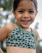 Ein junges Mädchen lächelt in die Kamera. Es trägt den grün-schwarz gemusterten Nalu Mädchen Bikini von Main Design (UPF 50+) und steht im Freien mit unscharfen Bäumen im Hintergrund.