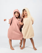 Zwei lächelnde Kinder stehen barfuß auf einem einfarbigen Hintergrund, gekleidet in den kuscheligen Main Design Kinder Badeponcho Flow. Das eine trägt Rosa, das andere Beige, aus weichem Musselin aus OEKO-TEX zertifizierter Baumwolle. Mit umeinander geschlungenen Armen nehmen sie eine verspielte Pose ein.