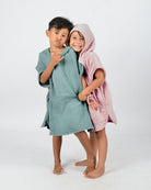 Zwei Kinder in dem Kinder Badeponcho Flow von Main Design lächeln verspielt. Die grünen und rosafarbenen OEKO-TEX-Baumwollponchos halten sie kuschelig warm, während sie barfuß stehen, einer macht ein lustiges Gesicht, der andere umarmt sie von hinten vor einem schlichten weißen Hintergrund.