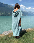 Eine Person mit langen Haaren, die einen hellblauen „Kinder Badeponcho Flow“ von Main Design trägt, steht mit dem Rücken zur Kamera auf einer Wiese am See, im Hintergrund sind Berge und ein teilweise bewölkter Himmel zu sehen.