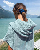 Ein Mädchen mit braunen Haaren, die zu einem Pferdeschwanz zusammengebunden sind, und einem blauen Haargummi steht an einem See und trägt den „Main Design Kinder Badeponcho Flow“. Strukturierter hellblauer OEKO-TEX-Baumwollstoff. Im Hintergrund sind Berge und ein blauer Himmel zu sehen.