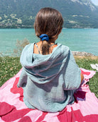 Eine Person mit braunen Haaren und einem blauen Haargummi sitzt auf einem rosa-roten Handtuch am Ufer eines Sees. Sie trägt den hellblauen „Main Design Kinder Badeponcho Flow“ aus OEKO-TEX-zertifizierter Baumwolle und blickt auf das Wasser und die fernen Berge.