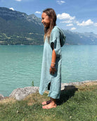 Ein junges Mädchen im „Main Design Kinder Badeponcho Flow“ steht barfuß auf dem Gras am Ufer eines türkisfarbenen Sees und lächelt, während im Hintergrund Berge und ein teilweise bewölkter Himmel zu sehen sind.