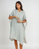 Eine Frau steht vor einem schlichten, hellen Hintergrund und trägt den Shoreline Breeze Poncho von Main Design - einen locker sitzenden, hellgrauen Baumwollkaftan mit kurzen Ärmeln und einem asymmetrischen Saum - und schaut ruhig in die Kamera.