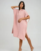 Eine Frau posiert barfuß in einem locker sitzenden, hellrosa Shoreline Breeze Poncho von Main Design mit kurzen Ärmeln und Seitenschlitzen. Der Poncho aus Baumwolle in Einheitsgröße bietet entspannten Komfort vor einem schlichten, hellen Hintergrund.