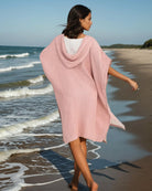 Eine Frau in einem hellrosa Main Design Shoreline Breeze Poncho läuft barfuß an einem Sandstrand unter einem klaren Himmel entlang. Dieser gemütliche Baumwollponcho in Einheitsgröße ist perfekt für entspannte Strandtage.