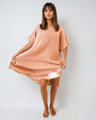 Eine Frau steht barfuß vor einem schlichten, hellen Hintergrund und trägt einen lockeren, strukturierten, hellpfirsichfarbenen Shoreline Breeze Poncho von Main Design in One Size mit kurzen Ärmeln, der die Seiten des Kleides leicht nach außen hält.