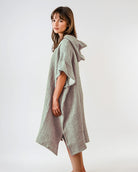 Eine Frau steht seitlich in dem Main Design Shoreline Breeze Poncho, einem lockeren, strukturierten hellgrauen Kleidungsstück mit kurzen Ärmeln und knielangem Saum in Einheitsgröße, das vor einem schlichten weißen Hintergrund gestylt ist.