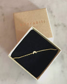 Das Armband von Adorabili, handgefertigt und 24K vergoldet mit Regenbogenanhänger, liegt in einer schwarzen Samtschachtel. Der braune Kartondeckel mit "Adorabili Paris" steht auf einer Marmorfläche dahinter.