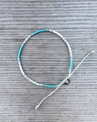 Das "Mama und Mini Armband Wave" von Main Design mit türkisfarbenen, silbernen und weißen Perlen sowie verstellbarem Verschluss wird auf grauer Oberfläche präsentiert - ideal als nachhaltiger Schmuck.