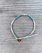 Das Mama und Mini Armband Wave von Main Design ist ein handgefertigtes Schmuckstück mit türkis, weißen und goldenen Perlen sowie einem kleinen runden Checkmark-Charm - nachhaltig präsentiert auf grauer Struktur.