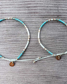 Zwei Main Design Mama und Mini Armband Wave Armbänder liegen nebeneinander auf einer grauen Oberfläche, mit blauen, weißen und goldenen Perlen und einem braunen runden Anhänger für nachhaltigen Schmuck.