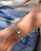 Nahaufnahme eines Handgelenks, das das Mama und Mini Armband Wave von Main Design trägt. Es ist mit tealfarbenen und silbernen Perlen sowie einem kleinen silbernen Herzanhänger versehen und wird zu einer Blue Denim-Jeans und einem hellen Top getragen.