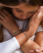 Ein kleines Mädchen mit geschlossenen Augen wird durch die Umarmung eines Erwachsenen getröstet. Beide tragen das bunte Mama und Mini Armband Wave von Main Design und fangen einen Moment der Wärme und Beruhigung mit nachhaltigem Schmuck ein.