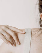 Nahaufnahme einer Hand, die sanft auf einer Schulter ruht und den Ring von Adorabili und Atelier Mouti by Adorabili trägt. Die Person ist mit einem leichten, schimmernden Stoff vor einem weichen, neutralen Hintergrund bekleidet.