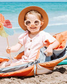 Ein lächelndes Kind sitzt auf einem bunten Strandstuhl am Meer und trägt einen Strohhut und eine runde Sonnenbrille. Das Kind hält ein buntes Windrad in einer Hand und sitzt bequem auf der Spielmatte und Krabbeldecke Boot des Hauptdesigns. Der Hintergrund zeigt blauen Himmel und Wellen.