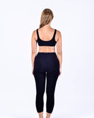 Eine Frau steht barfuß und blickt von der Kamera weg vor einem weißen Hintergrund. Sie trägt ein schwarzes ärmelloses Fiji Active Top von Main Design aus Lenzing™ Ecovero™ und hoch taillierte schwarze Leggings. Ihr glattes, blondes Haar fällt ihr in diesem nachhaltigen Loungewear-Set über den Rücken.