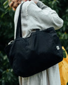 Eine Person in einem hellen, strukturierten Mantel trägt die "Stofftasche klein von OMOM" von Main Design, eine kleine schwarze Tasche mit einem weißen Anhänger, während sie im Freien vor grünem Laub steht.