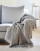 Die Decke Savanna von Main Design mit grauem Leopardenmuster und fransigen Rändern wird in einem modernen Wohnzimmer neben einem kleinen runden Holztisch über die Armlehnen und die Sitzfläche eines hellgrauen Polstersessels drapiert.